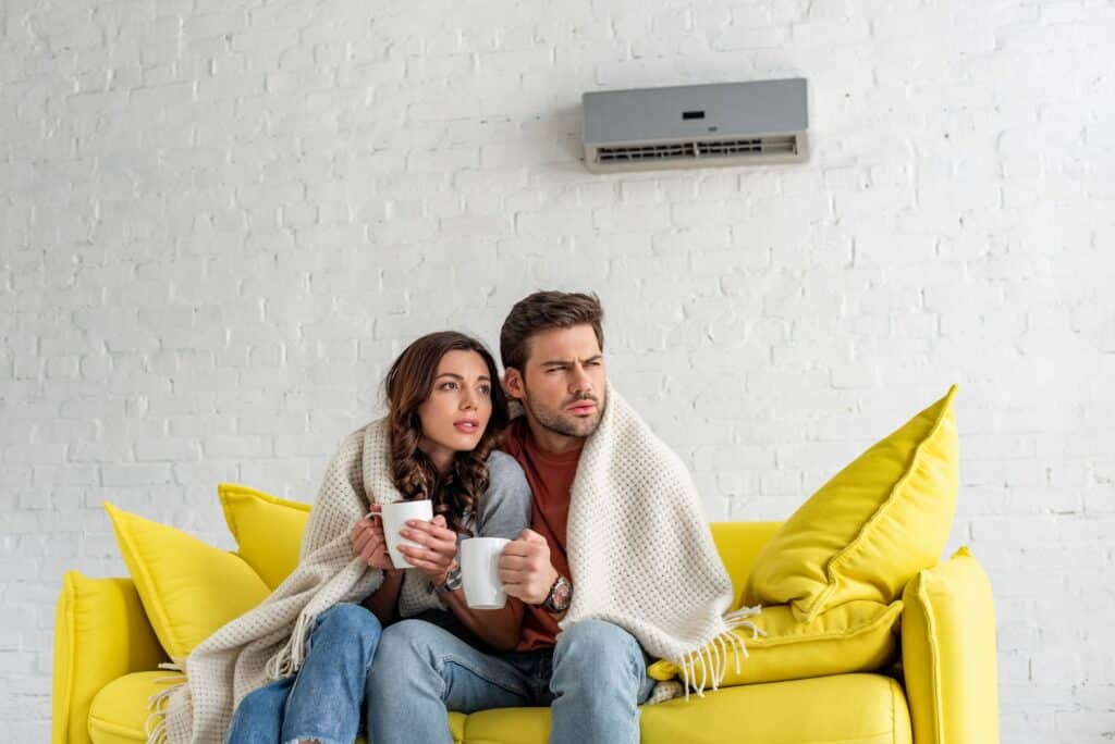 frozen couple with cups warming under blanket while sitting under air conditioner at home