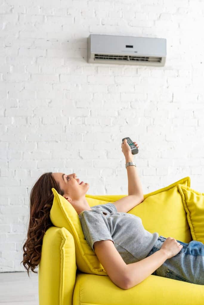 pretty young woman lying on yellow sofa under air conditioner and holding remote control