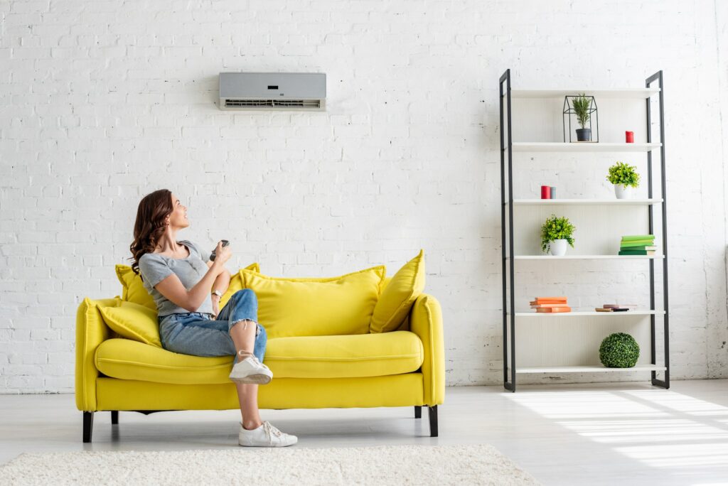 pretty young woman sitting on yellow sofa under air conditioner at home