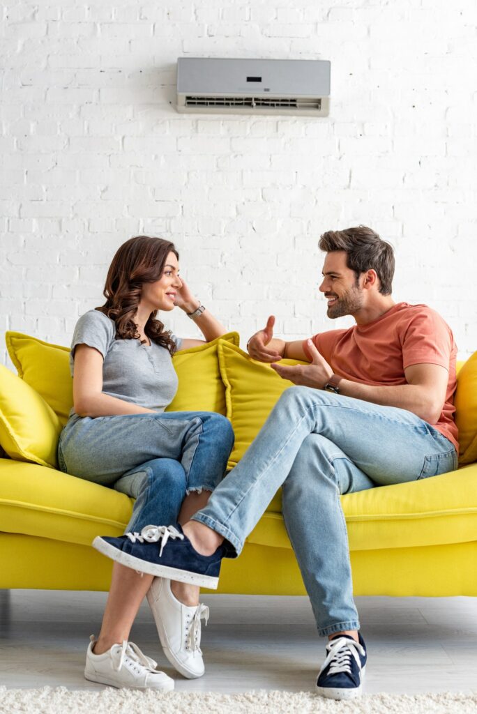 smiling man and woman sitting on yellow sofa under air conditioner at home